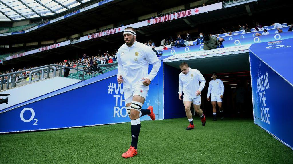 Lewis Ludlow runs out of the tunnel at Twickenham wearing a white England branded jacket and white shorts