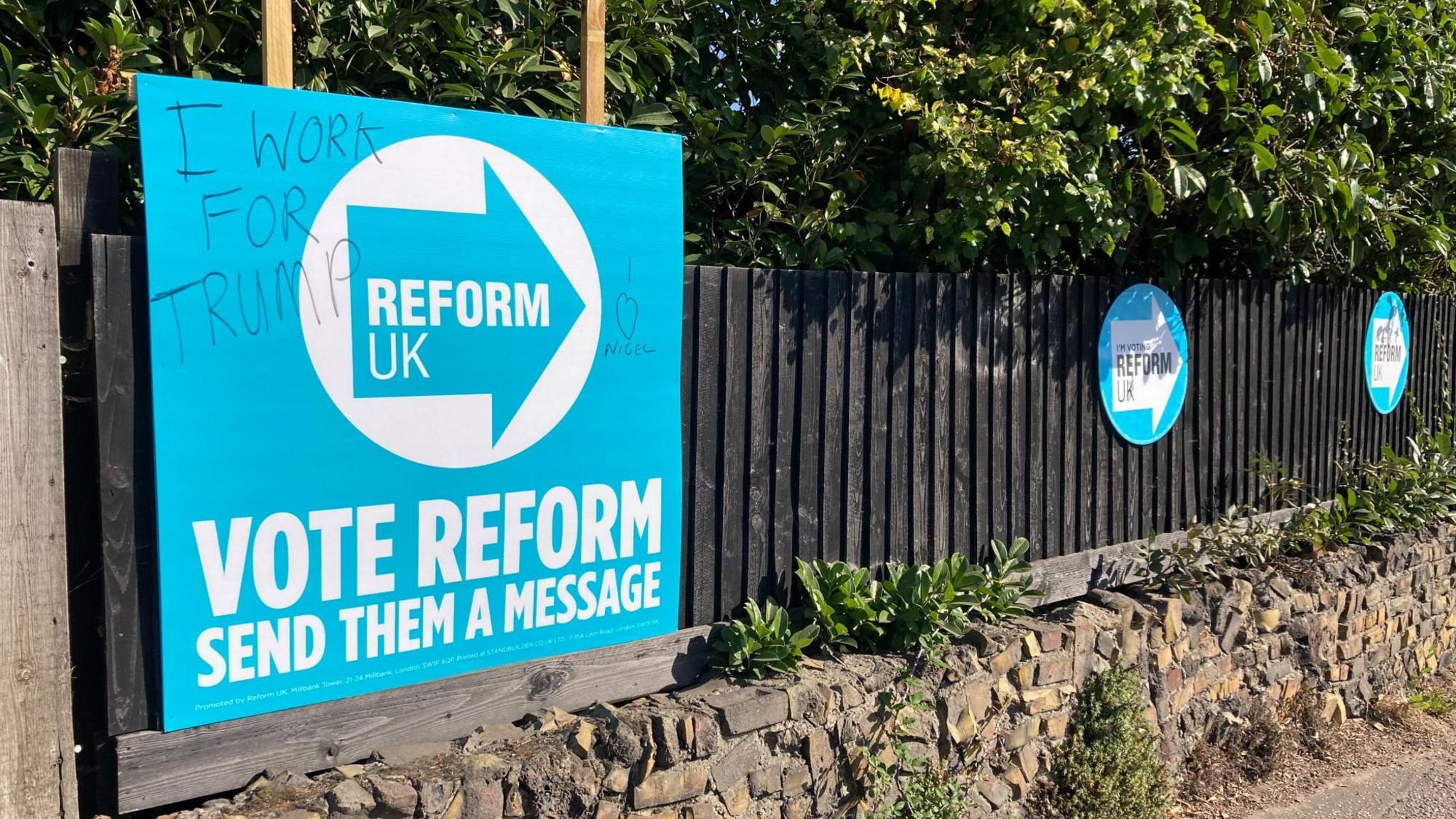 A rectangular blue and white "VOTE REFORM" placard stuck on a wooden fence above a brick wall on a pavement. It has graffiti in black marker pen saying "I WORK FOR TRUMP" AND "I [heart-shape] NIGEL" on it. There are two smaller, round "I'M VOTING REFORM UK" placards further along the fence.