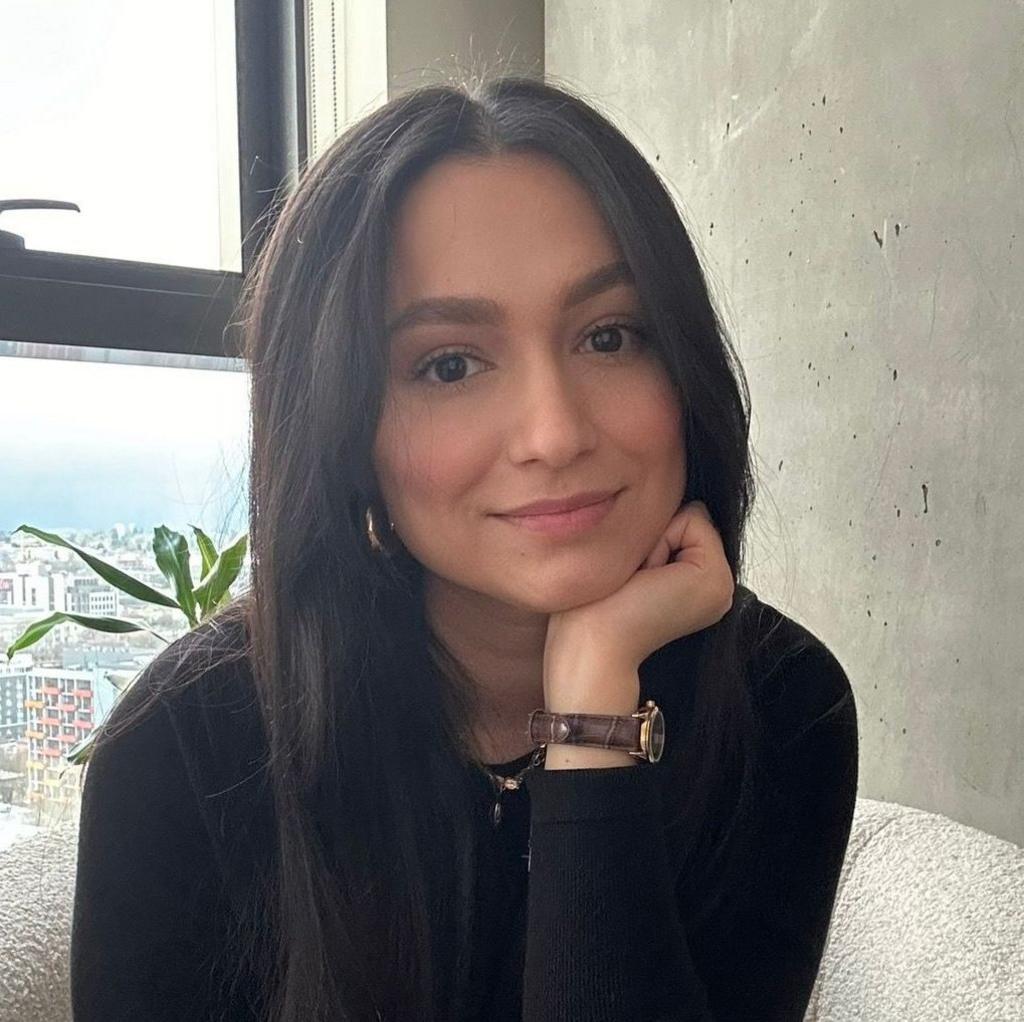 Person with long dark hair seated in a textured upholstered chair in the foreground, wearing a black long-sleeved top and a wristwatch. One hand supports the chin while facing the camera. A large window and a potted plant are visible to one side, with buildings seen through the window. A plain concrete wall forms the background.
