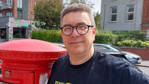 A man standing beside a red post box. He has short grey hair and is wearing glasses and a navy t-shirt.