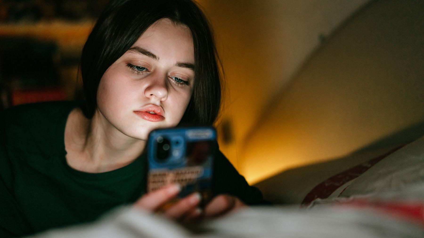 A teenage girl looks at her phone at nigh ttime in low lighting with the glow of a lamp in the background. The light from the phone lights her face