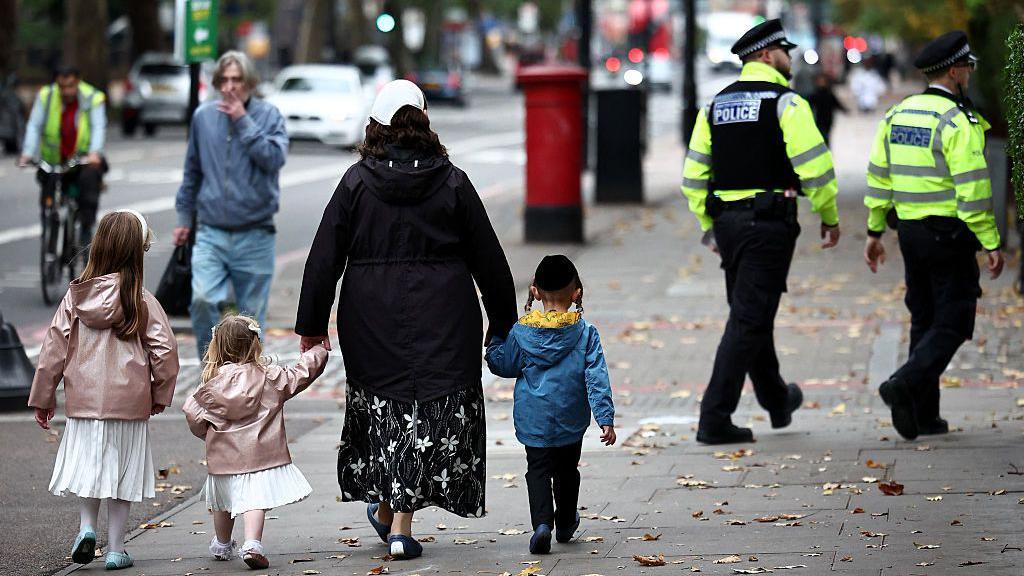 A family including two young girls in white dresses and pink coats along with a little boy in a blue coat. They walk down the street with their backs to the camera and in the background two Metropolitan Police officers in uniform can be seen walking in the same direction.
