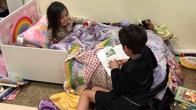 A young boy clad in black shorts and a black top sits on the ground, holding a book on the edge of his sister's bed as he reads it to her; she sits under a purple blanket, a rainbow visible on the headboard