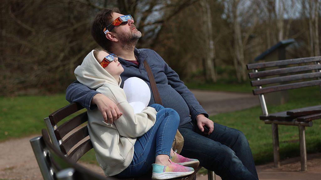 A man and a child watching a partial solar eclipse at Keele university on 29 March, 2025. They are wearing special glasses and are sat on a bench and there is part of what looks like an empty bench next to them. There is grass in the background.