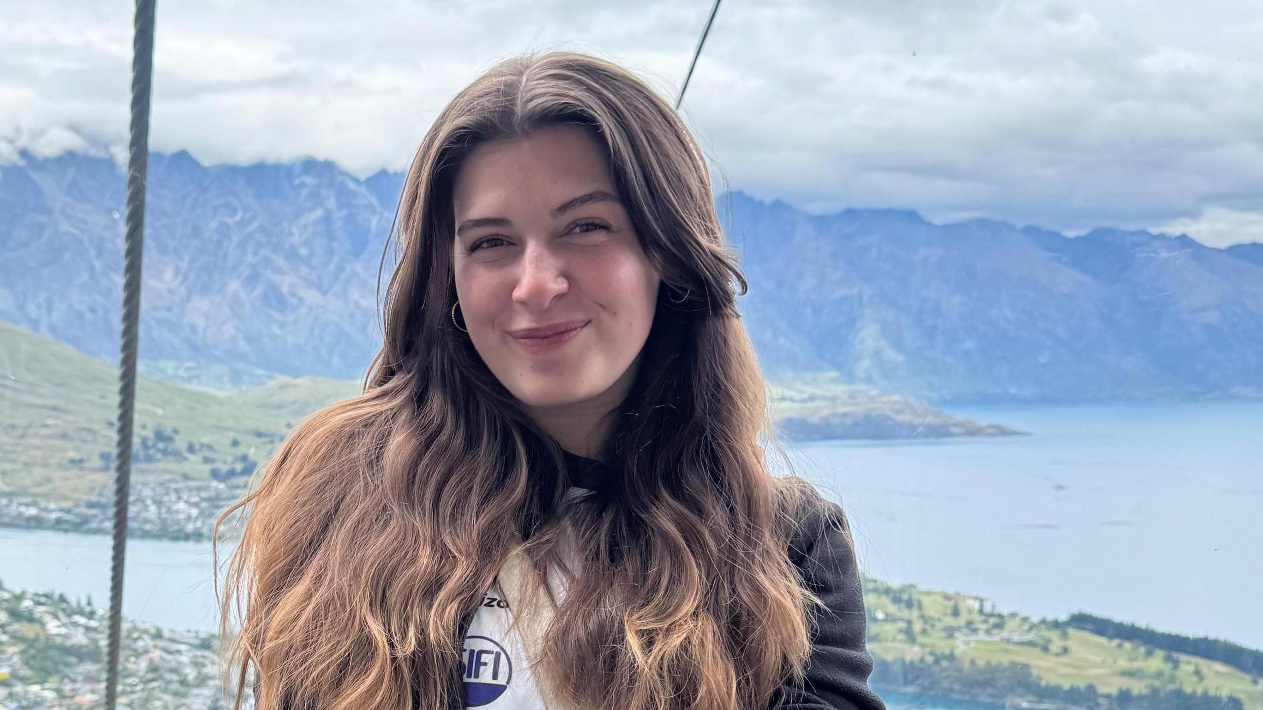 Elena is sitting in a cable car, with a view of a lake and mountains behind her. She is wearing a long top with the sleeves rolled up and has a cream handbag across her body. She has long brown hair and is smiling at the camera, with her hands crossed on her lap.