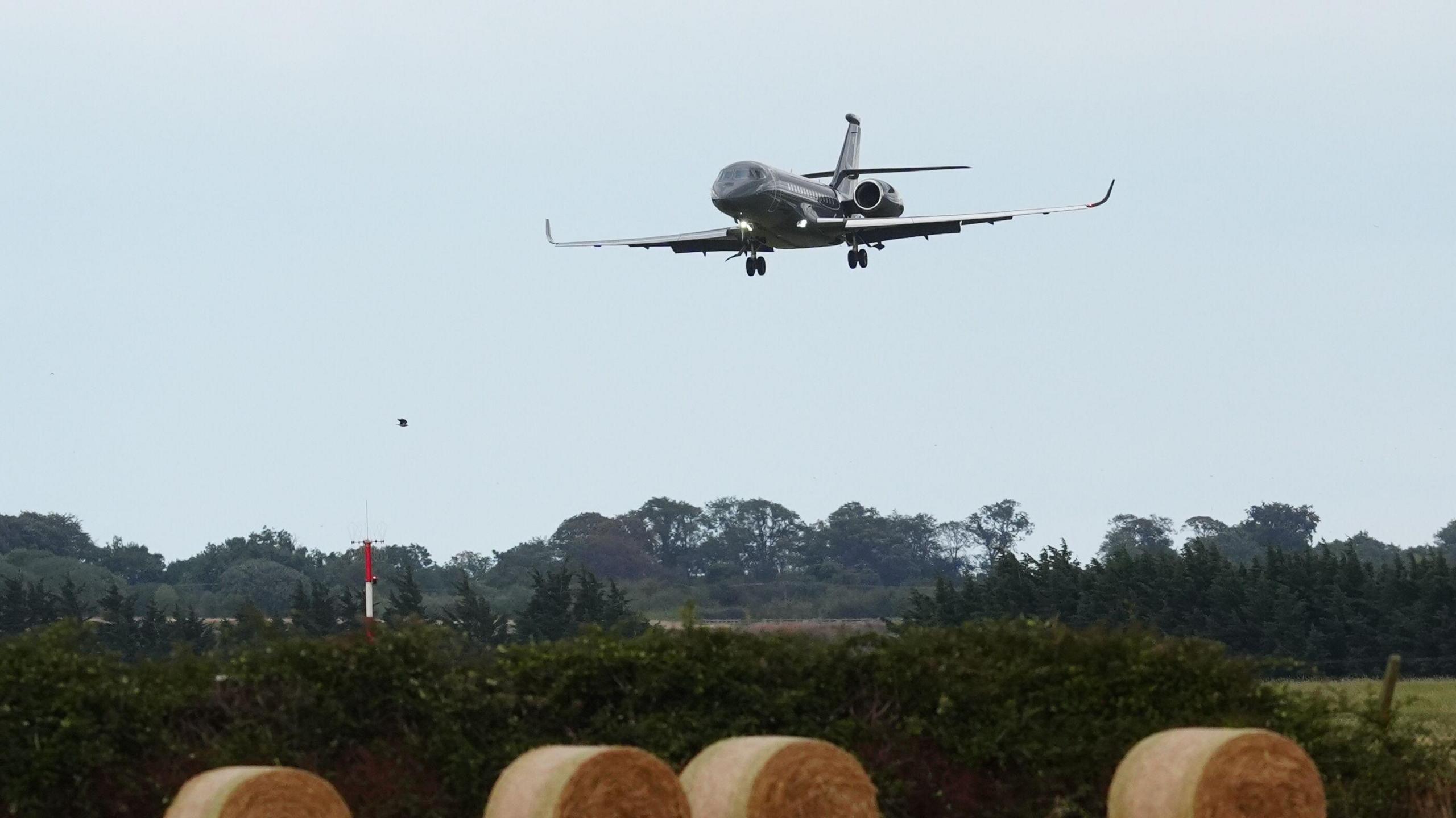 A dark grey plane comes into land near a field of hay bales and green trees, the sky behind the aircraft is blue.