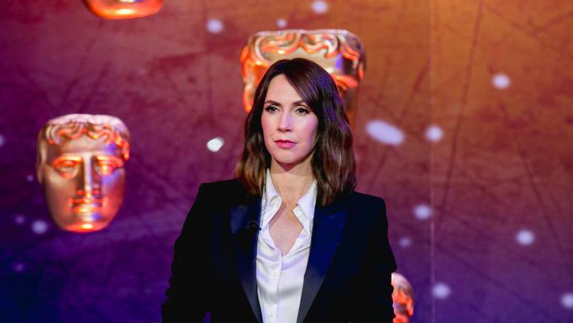 Alex jones on the bafta awards stage with the golden actors mask on in the backgrounbd. Alex is wearing a blazer and a white blouse.