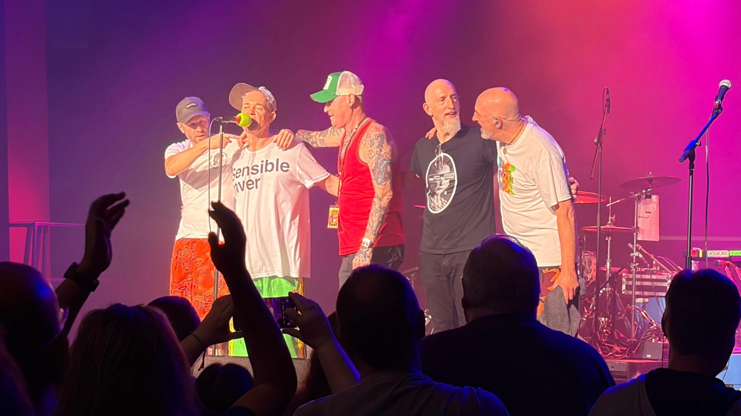 EMF - a group of five middle-aged male band mates - stand side-by-side on stage following a performance. They are dressed in streetwear and their instruments and microphones are illuminated in purple behind them. A crowd is watching on.