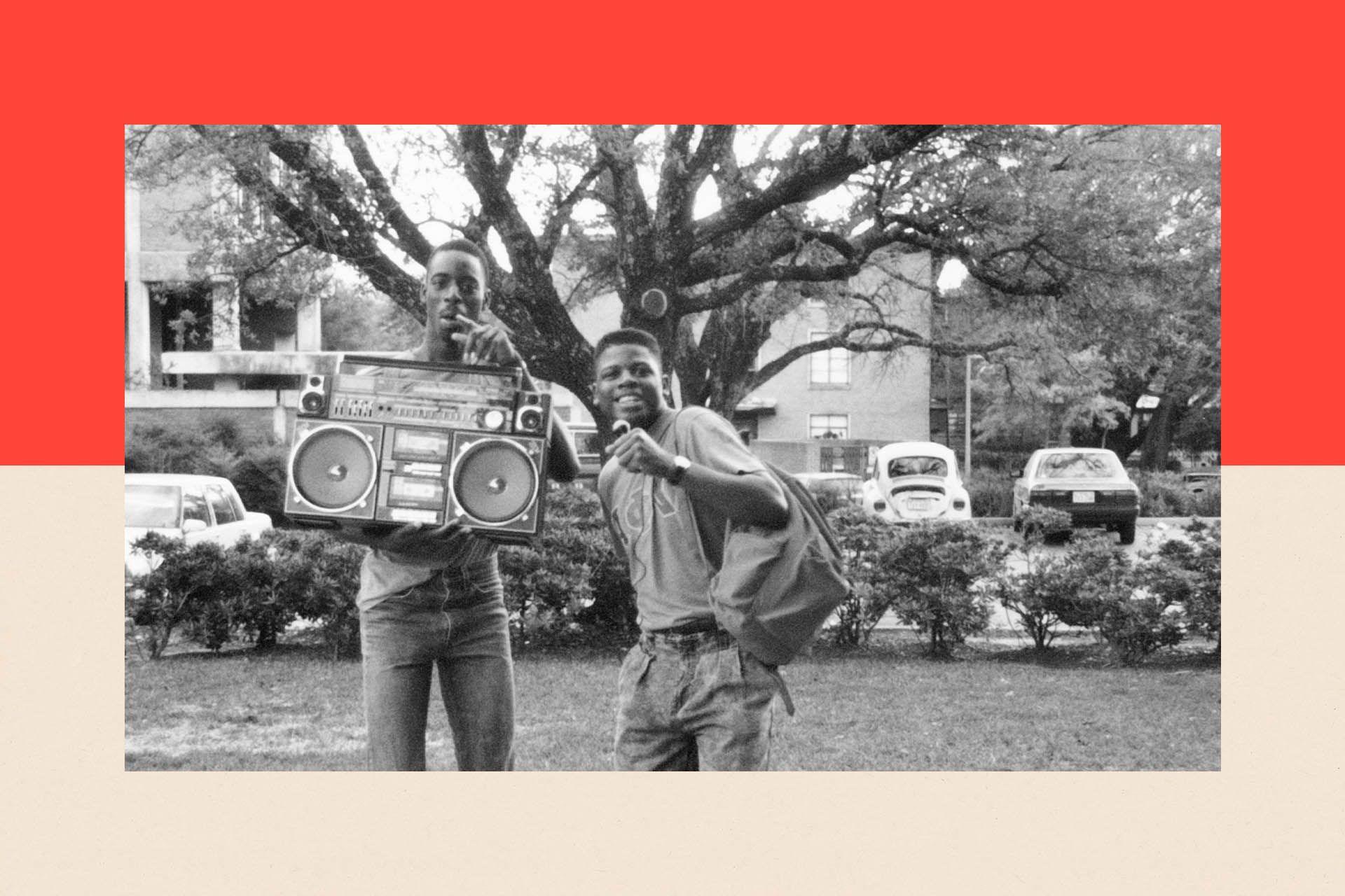 Two young men stand by a boombox and smile. The photograph is in black and white