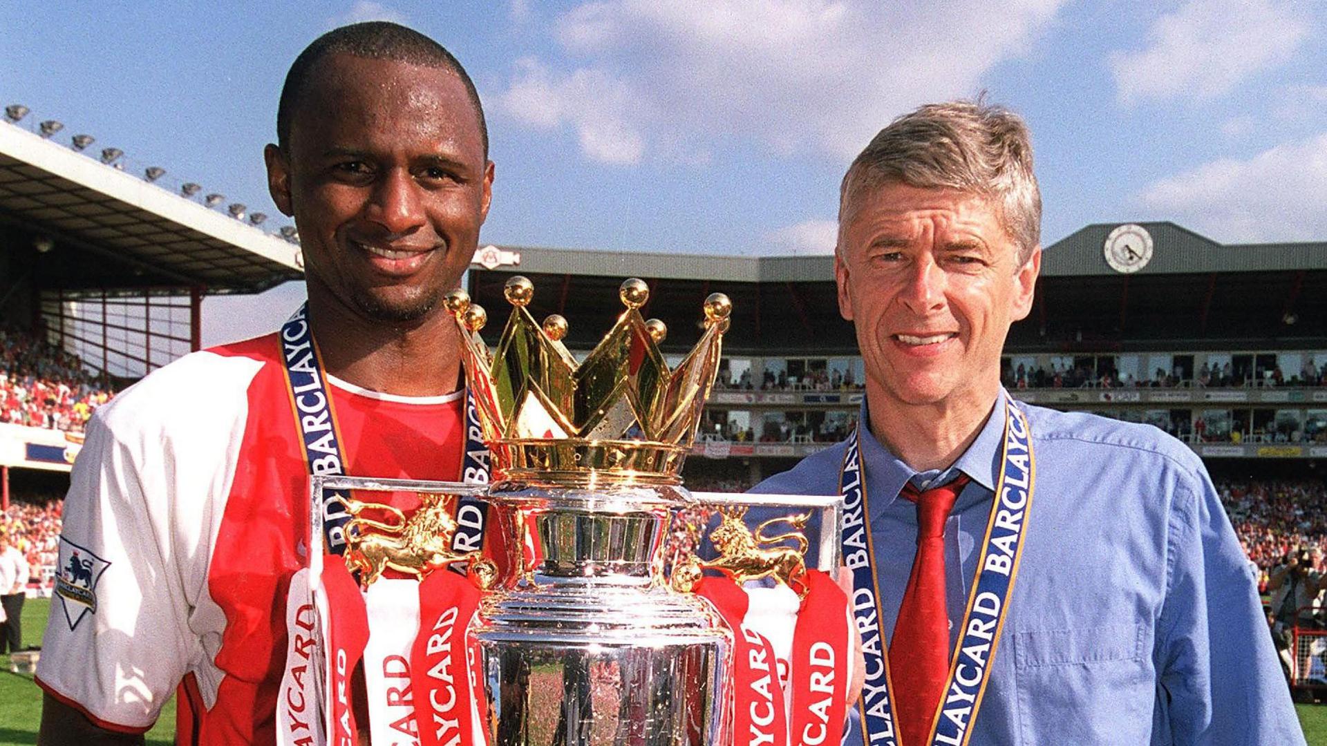 Patrick Vieira wears a red-and-white Arsenal shirt as he holds the Premier League trophy bearing red and white ribbons at chest height. He stands next to Arsene Wenger, who wears a blue shirt with a red tie. Both men, who are visible from chest up, have blue ribbons around their necks holding medals which are out of shot. In the far background the Clock End of Highbury Stadium can be seen