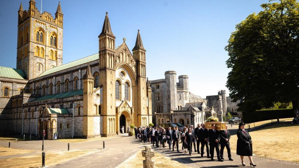 The funeral procession for Ann Widdecombe with Buckfast Abbey towering in the background against a blue sky backdrop.