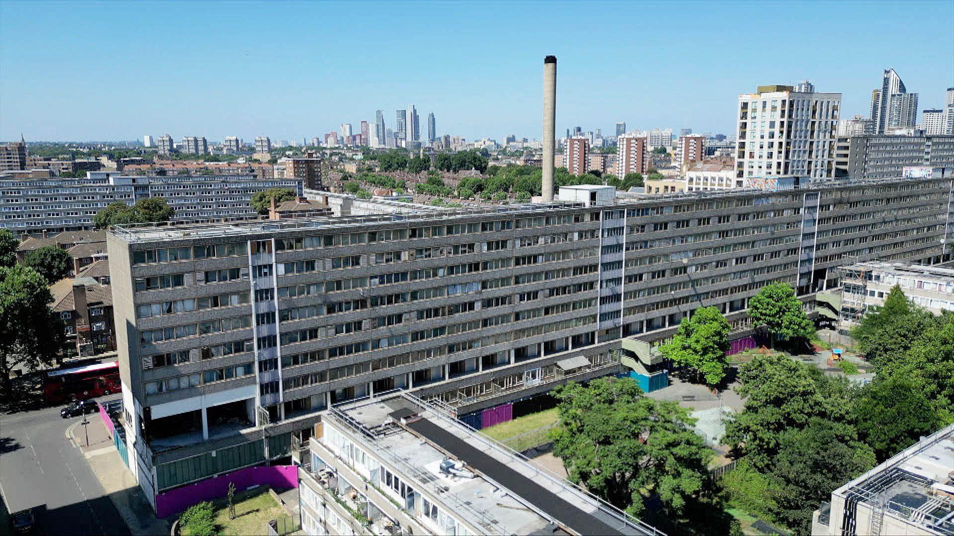 An aerial view of abandoned flats in south London. There are trees in bottom right of the frame and the city skyline can be seen in the distance against a blue sky.