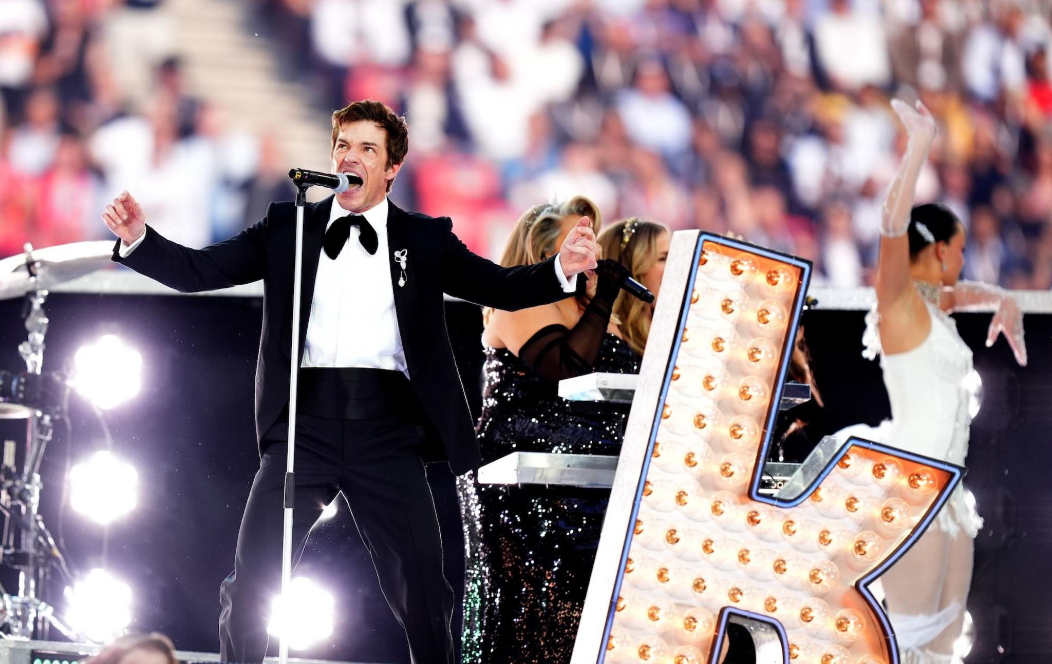 Brandon Flowers performs with The Killers at the  UEFA Champions League final. He's dressed in a tuxedo with a bow tie, standing next to a large billboard-style letter K.