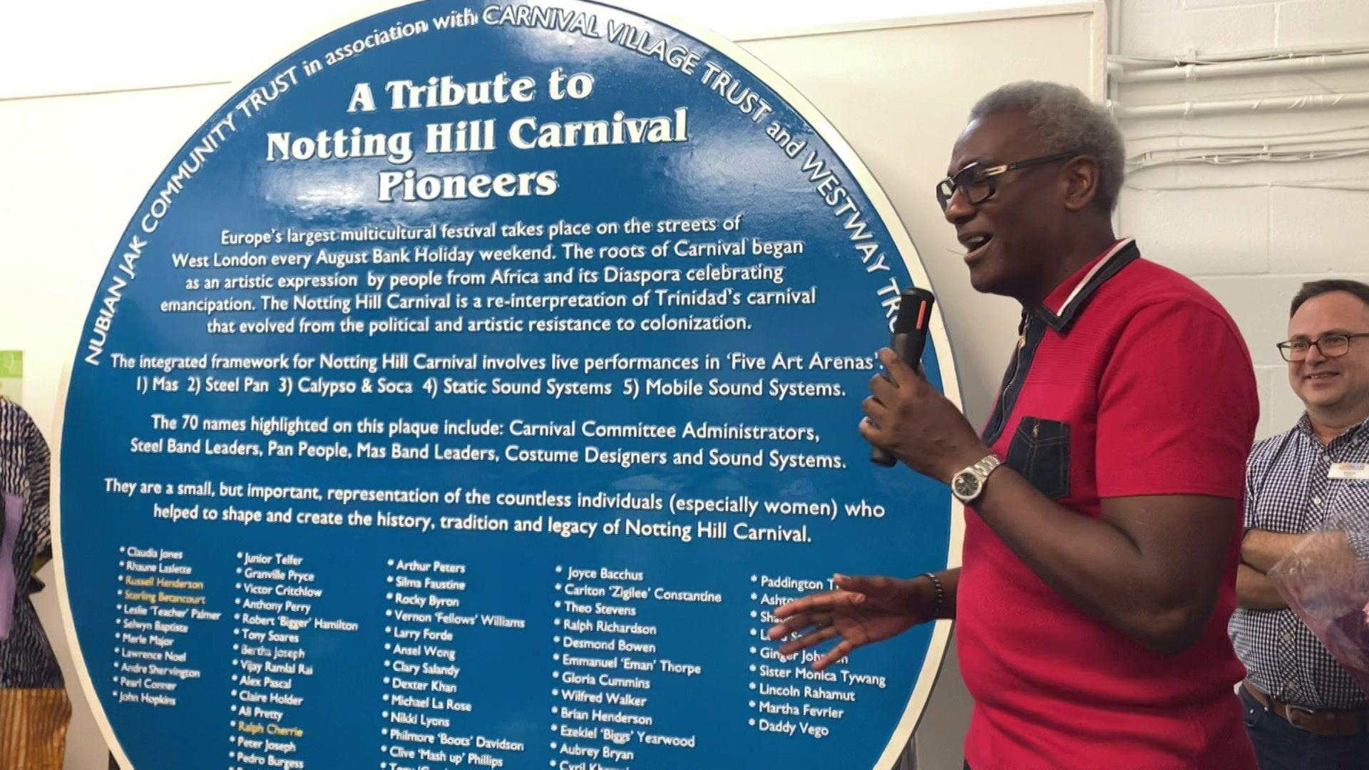Man in a red polo shirt holds a microphone in front of a large round blue plaque listing 70 carnival pioneers