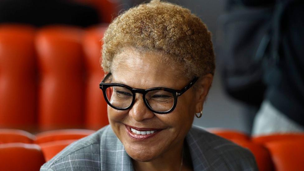 This is a head and shoulders pic of Karen Bass who is smiling broadly and wearing a grey, checked suit. The background, slightly out of focus, appears to be red seating