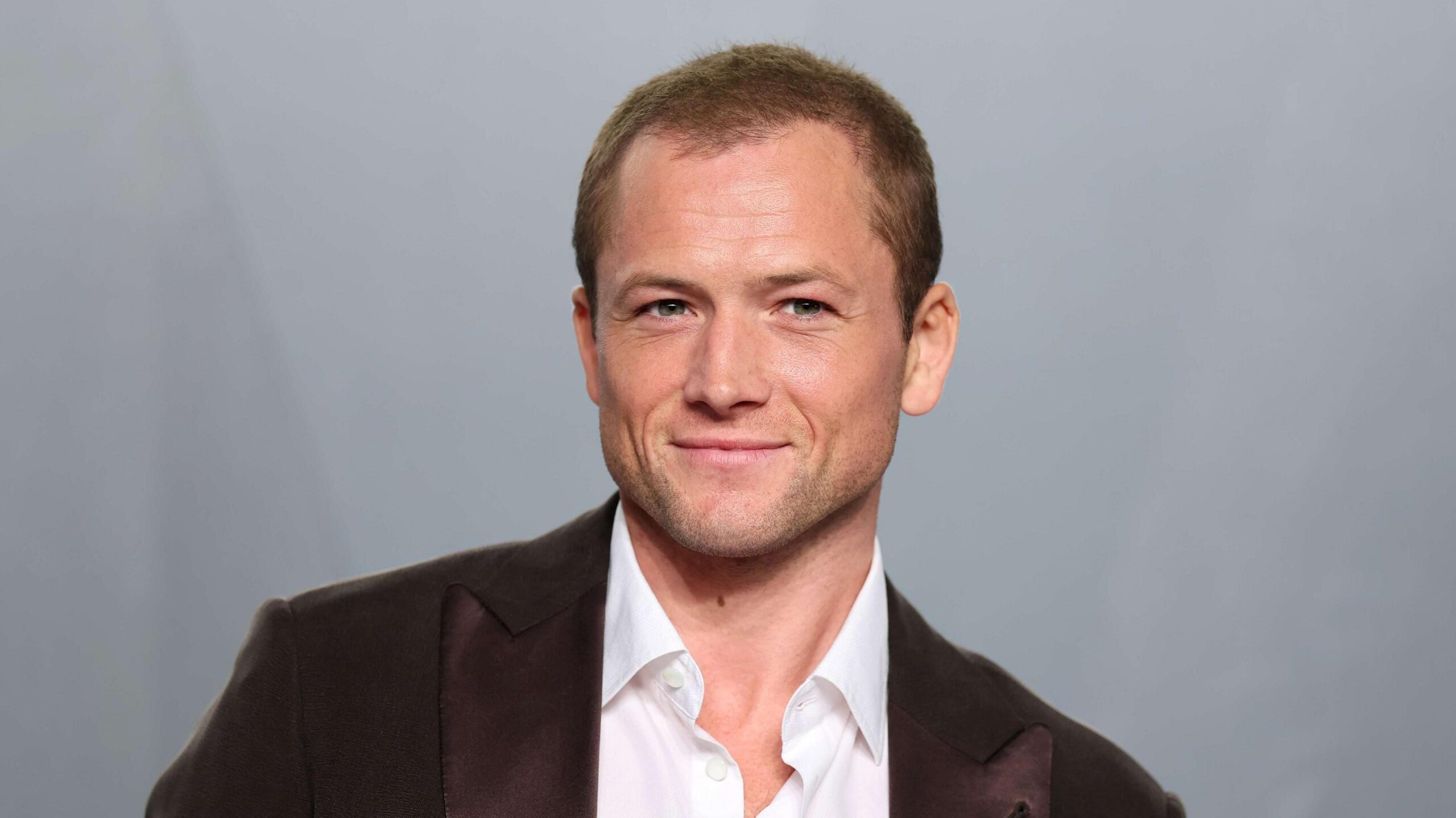 Egerton, a man in his late 30s wearing a white shirt and a brown suit jacket. There's a grey background behind him.