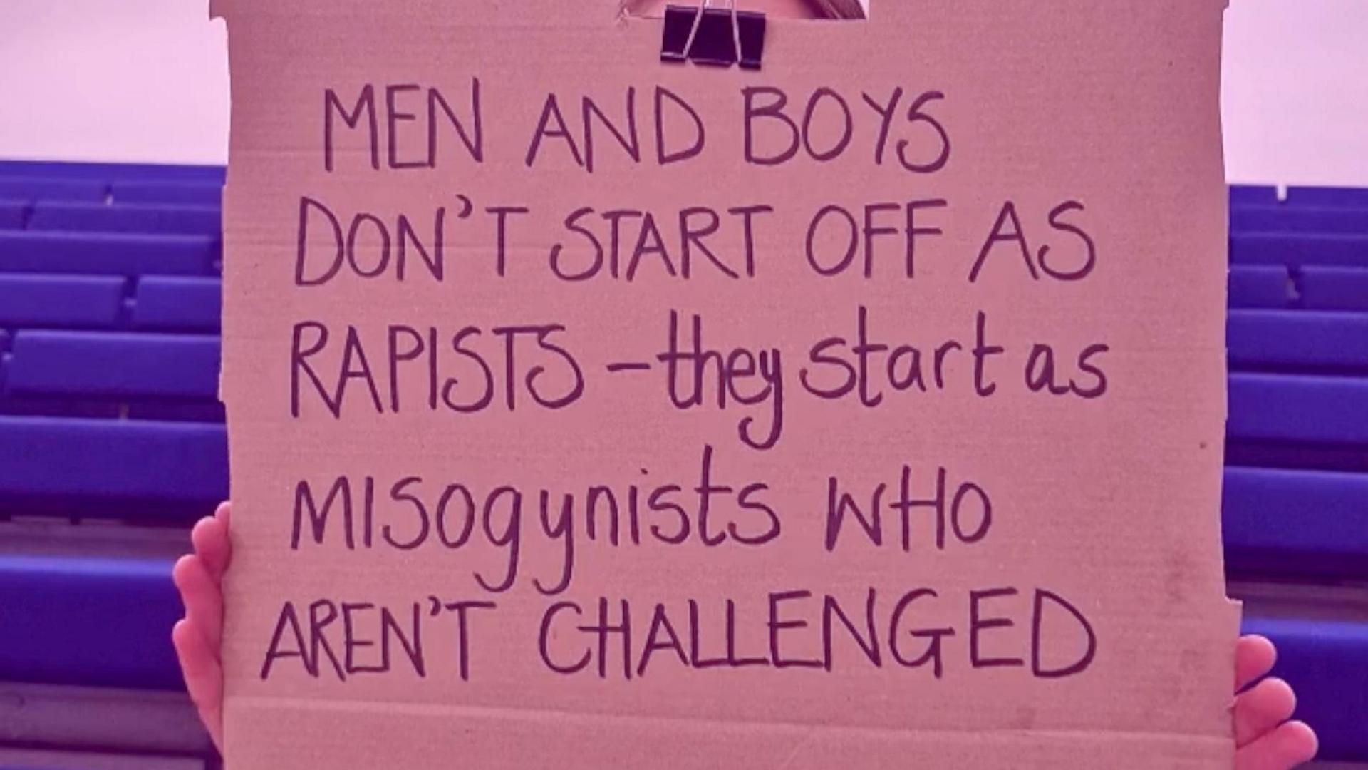 A cardboard sign is held in front of a girls face, her hands are visible on the edges. The phrase 'Rapists - they start as misogynists who aren't challenged' is written on the sign in black marker pen.
