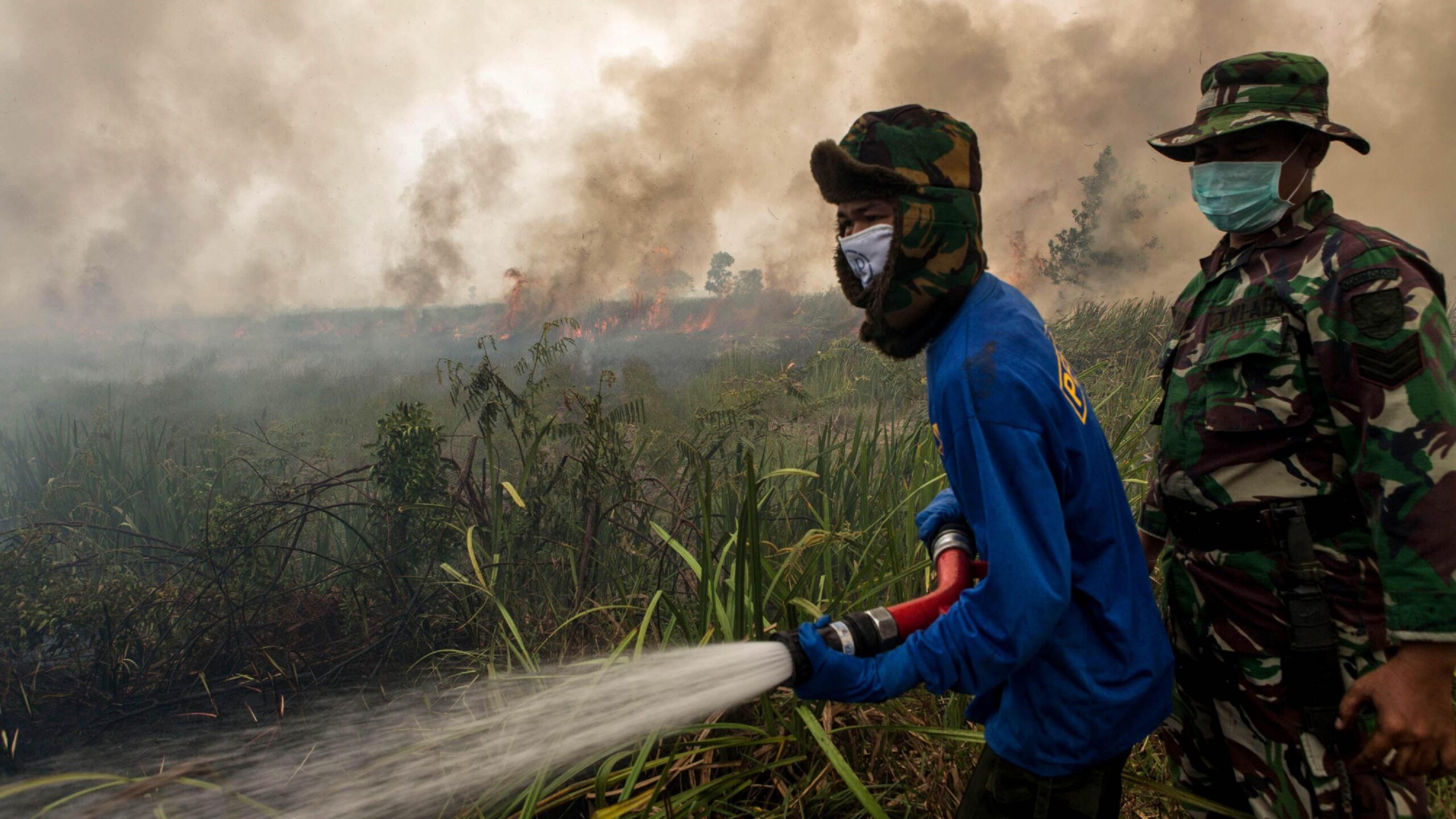 Two firefighters tackle a fire, with smoke and flames in the background. One man is dressed in a blue top; the other is in green camouflage kit. The man in blue is holding a water hose. Both are wearing masks.
