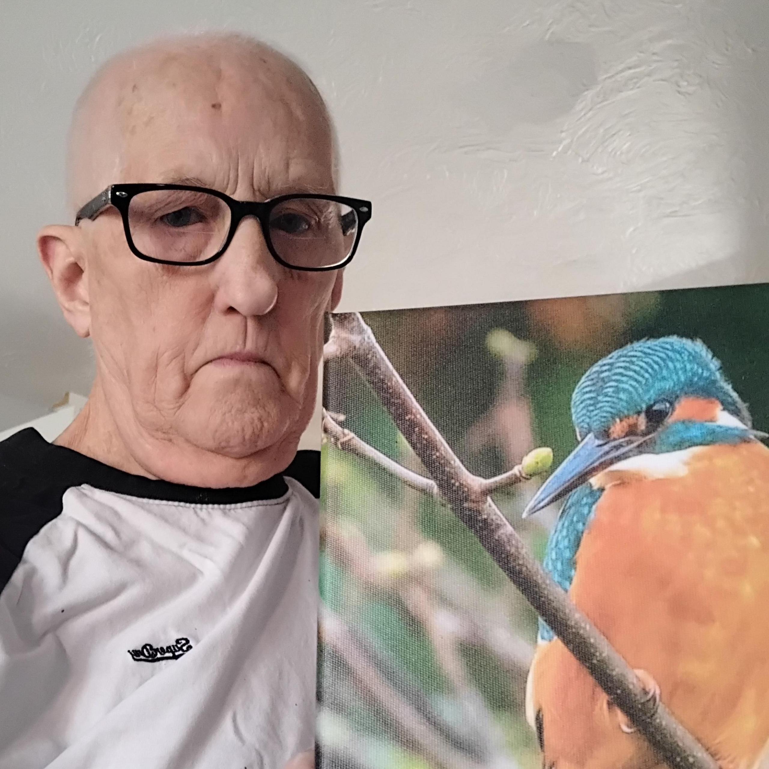 Chris Fell, a bald white man, wears a white t-shirt and glasses as he holds up a picture of a bird.