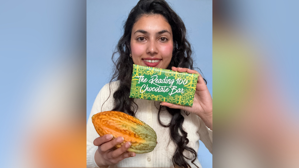 A woman with long brunette hair holding a large cocoa bean and a wrapped chocolate bar up to the camera while smiling.