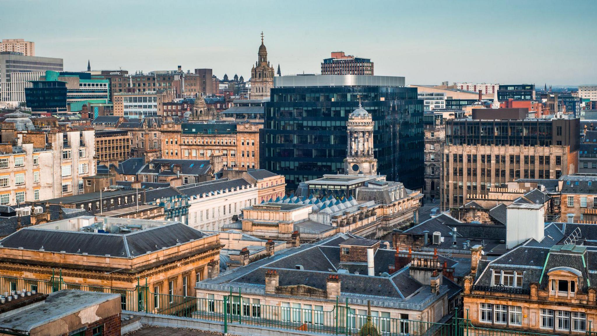 Panoramic view of a Glasgow city centre skyline featuring a mix of historic sandstone buildings and modern glass office blocks under a pale blue sky. Rooftops fill the foreground, while prominent church towers and civic buildings rise above the urban landscape in the distance.