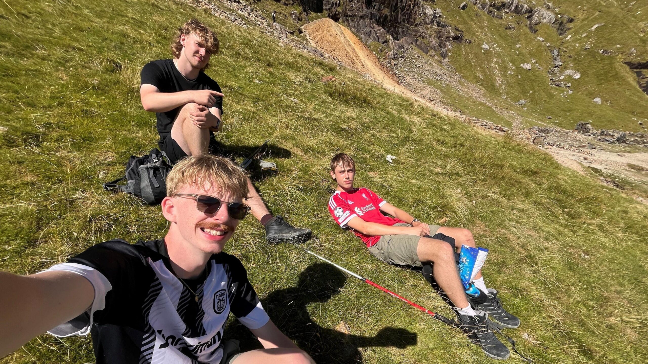 Three young men sit on a grassy hillside during a mountain walk. James Munday takes a selfie in the foreground wearing a black and white sports shirt and sunglasses. He has blonde hair and a moustache. Two others sit higher up the slope. Stan sits highest on the hill and has a blonde mullet and is wearing a black shirt and shorts.  Fin lays next to them wearing a red shirt and kaki shorts. He has short brown hair. Walking poles, backpacks and rocky mountain scenery can be seen around them.