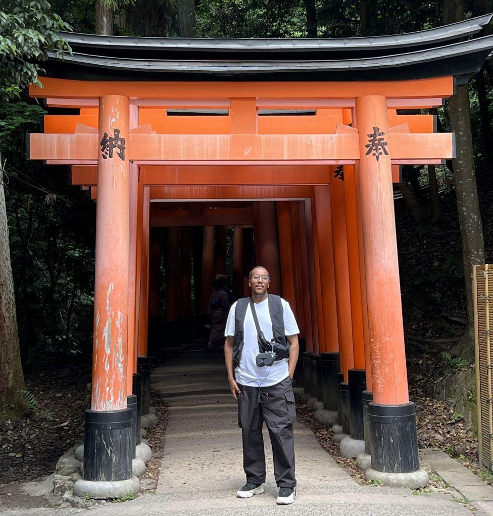 Aden Ibrahim, a man in white T-shirt and black trousers, stands under an orange torii gate in Japan