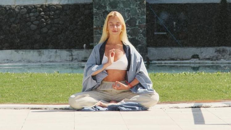 Anastasia sits cross-legged meditating in a garden on a sunny day