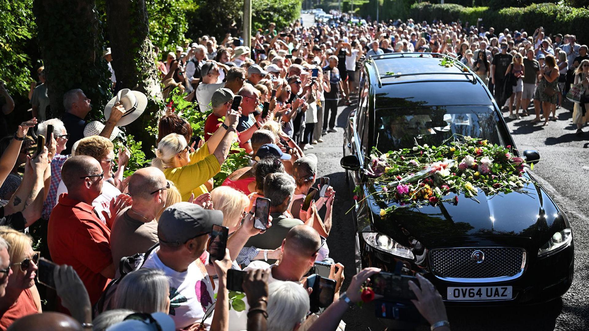 Bystanders flock towards a black hearse and throw flowers onto its bonnet as it drives through the town carrying the coffin of Bonnie Tyler.