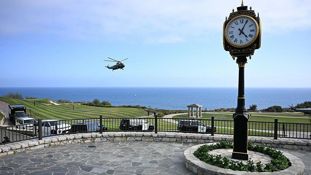Trump's White House helicopter seen landing at the California golf course on Tuesday. There is a clocktower in the foreground and golf greens and ocean in the background