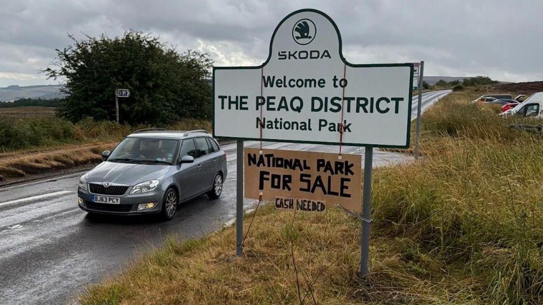 A road sign which reads SKODA WELCOME TO THE PEAQ DISTRICT NATIONAL PARK. A cardboard sign is hung around the larger sign which reads NATIONAL PARK FOR SALE CASH NEEDED.