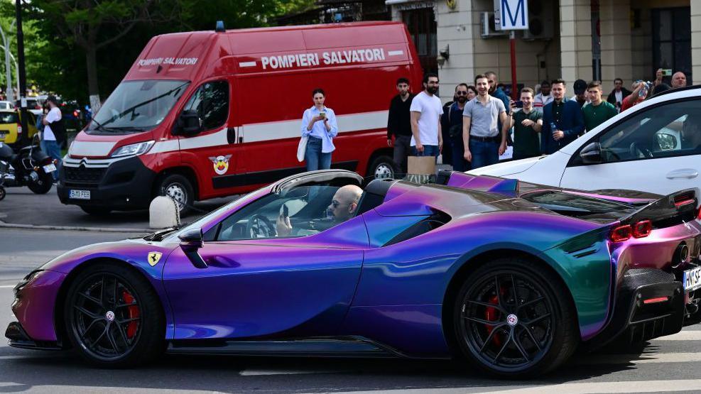 Andrew Tate driving a purple, open-top Ferrari with a crowd in the background