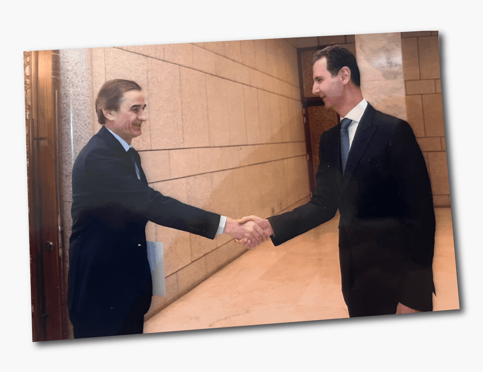 A photo of Hussam Luka shaking hands with Assad - both are wearing dark suits and smiling. They are pictured in a room with marble-looking walls and floor