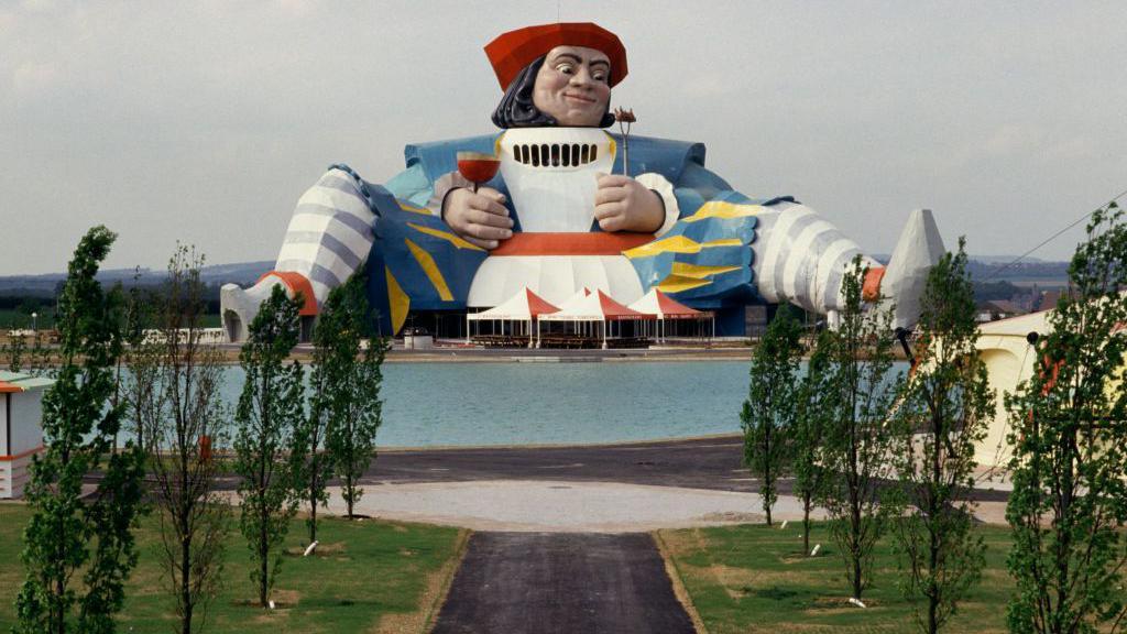 A giant statue of a man, dressed in medieval clothing and folding a fork in one hand and a glass of red wine in the other, sits with his legs spread above a lake and trees in the former theme park