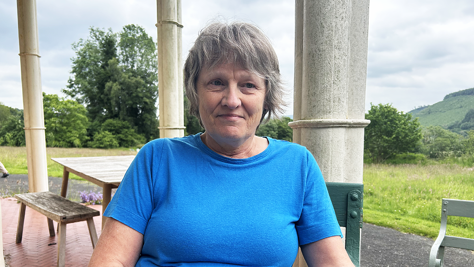 Jenny Kincova is a white woman in her 60s with feathered short light grey hair. She is wearing a sea-blue short-sleeved t-shirt. She is smiling slightly and looking just to the right of the camera. Behind her are pillars supporting an out of view covered veranda, and a wooden table and bench. She is seated. Beyond the veranda is a large grassy lawn which has been left to grow naturally in most areas and trees behind that. A hill is visible in the further distance.