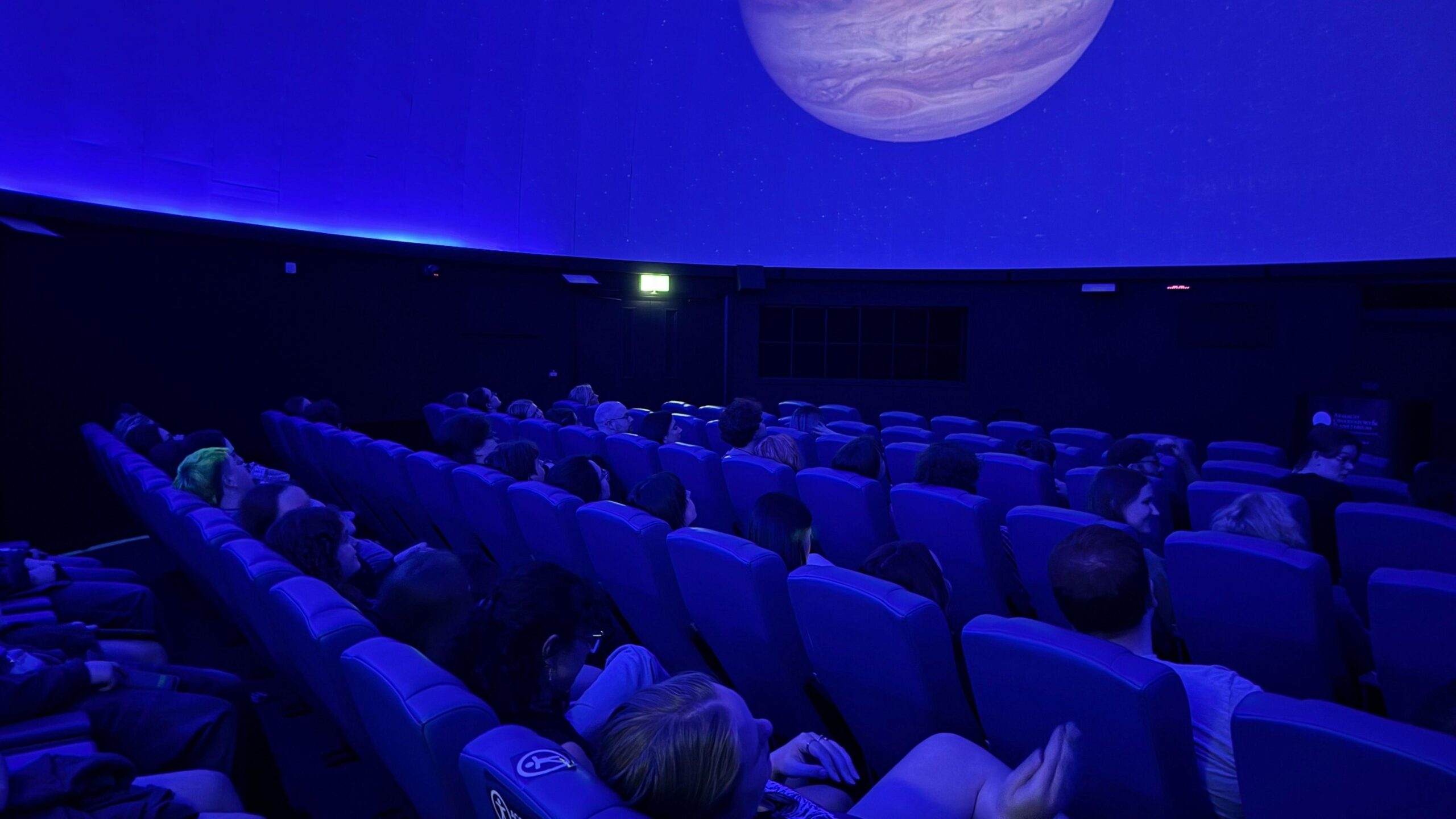 The audience sit in rows of seats in the planetarium's main dome room. The room is lit blue.