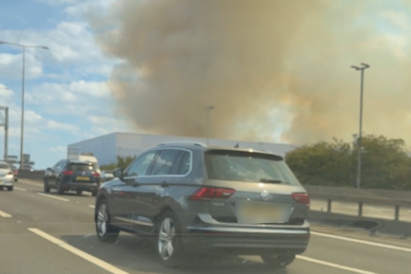 A silver family car is moving on a motorway. Other cars are in the frame, and in the background, large plumes of smoke rise from an industrial warehouse building.