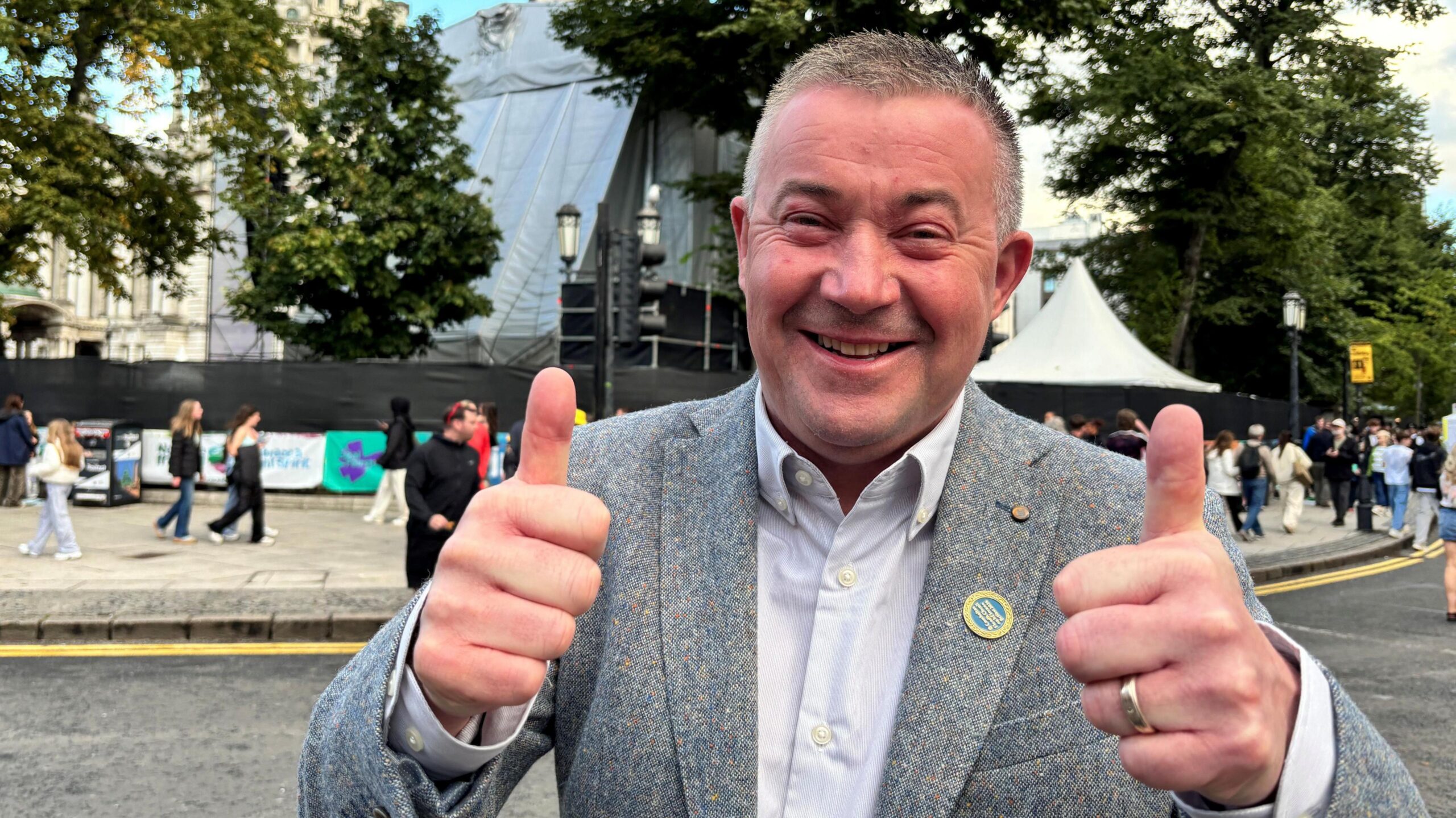 A man with grey/black hair smiling and giving thumbs up to the camera, he has a grey suit on
