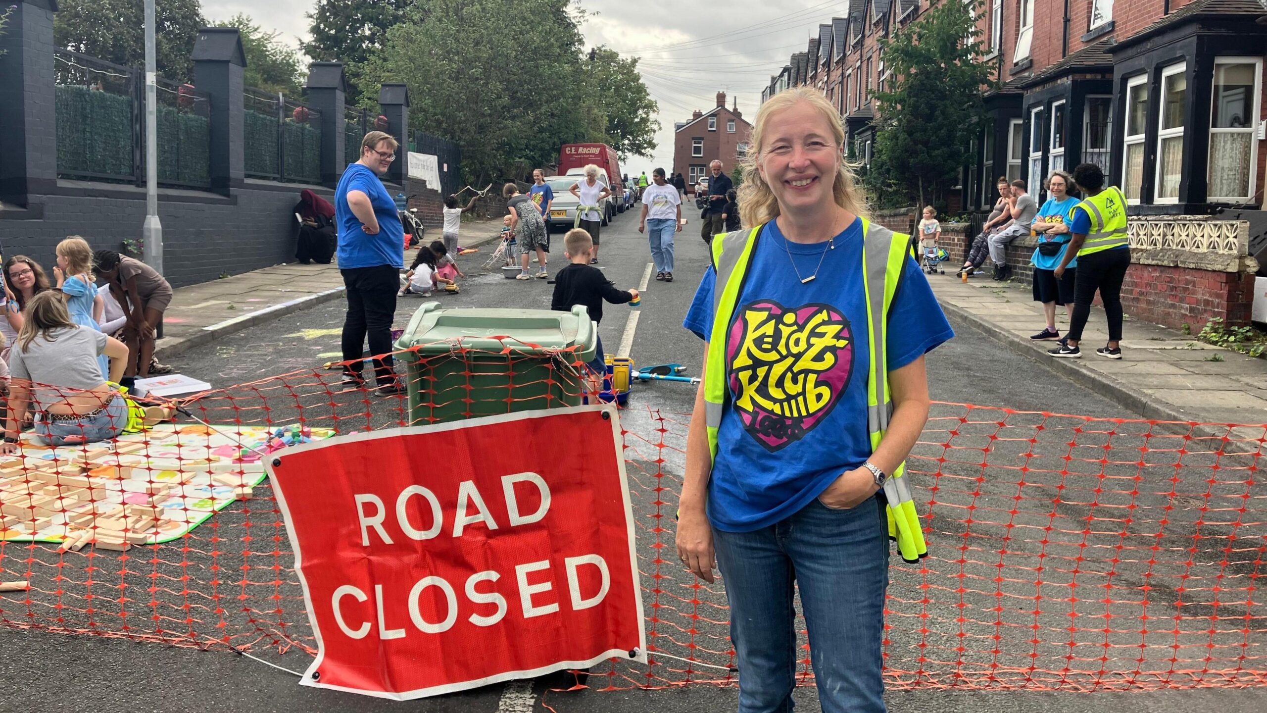 A white woman in her 50s with long blonde hair is wearing a blue t shirt and a high vis jacket. She is smiling at the camera and standing in the middle of a residential street which has been closed to traffic. Behind her children are playing with toys.