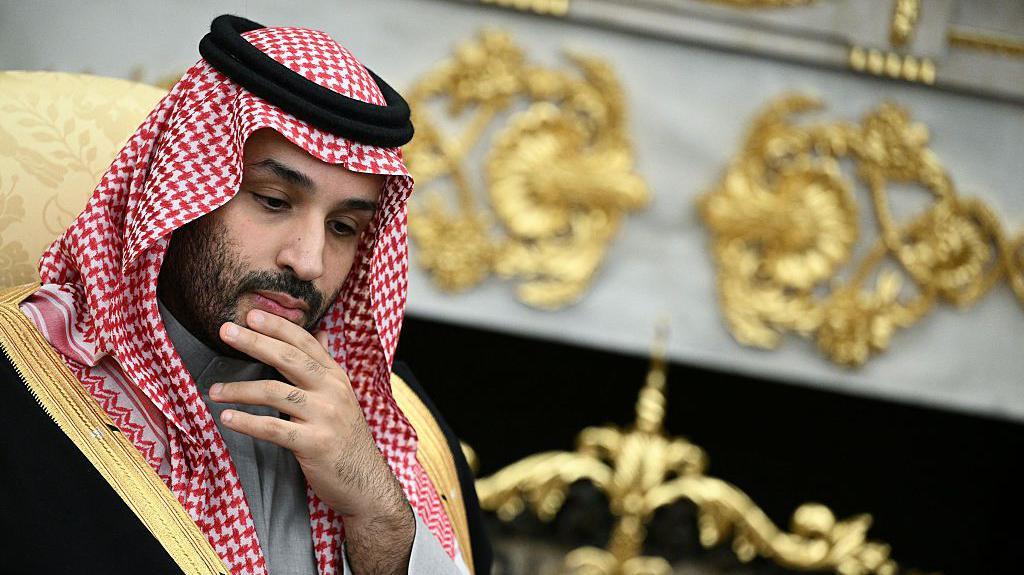 He is rubbing his chin, thinking. He is wearing traditional Arab clothing, with the red and white head dress. He is wearing a black robe with gold edges.