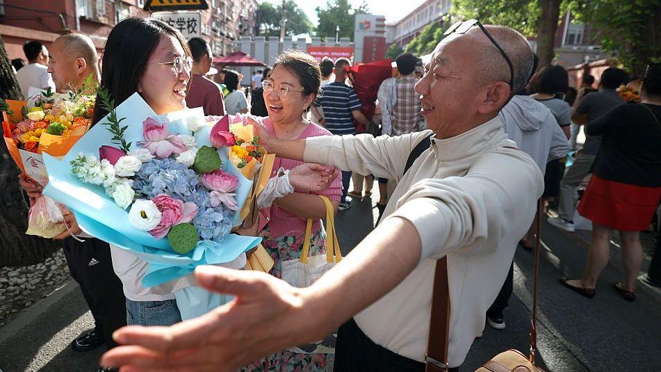 A student is embraced by his father outside the examination venue after the 2026 China's National College Entrance Exam on June 10, 2026 in Beijing, China. She is grinning and holding a bouquet of flowers.