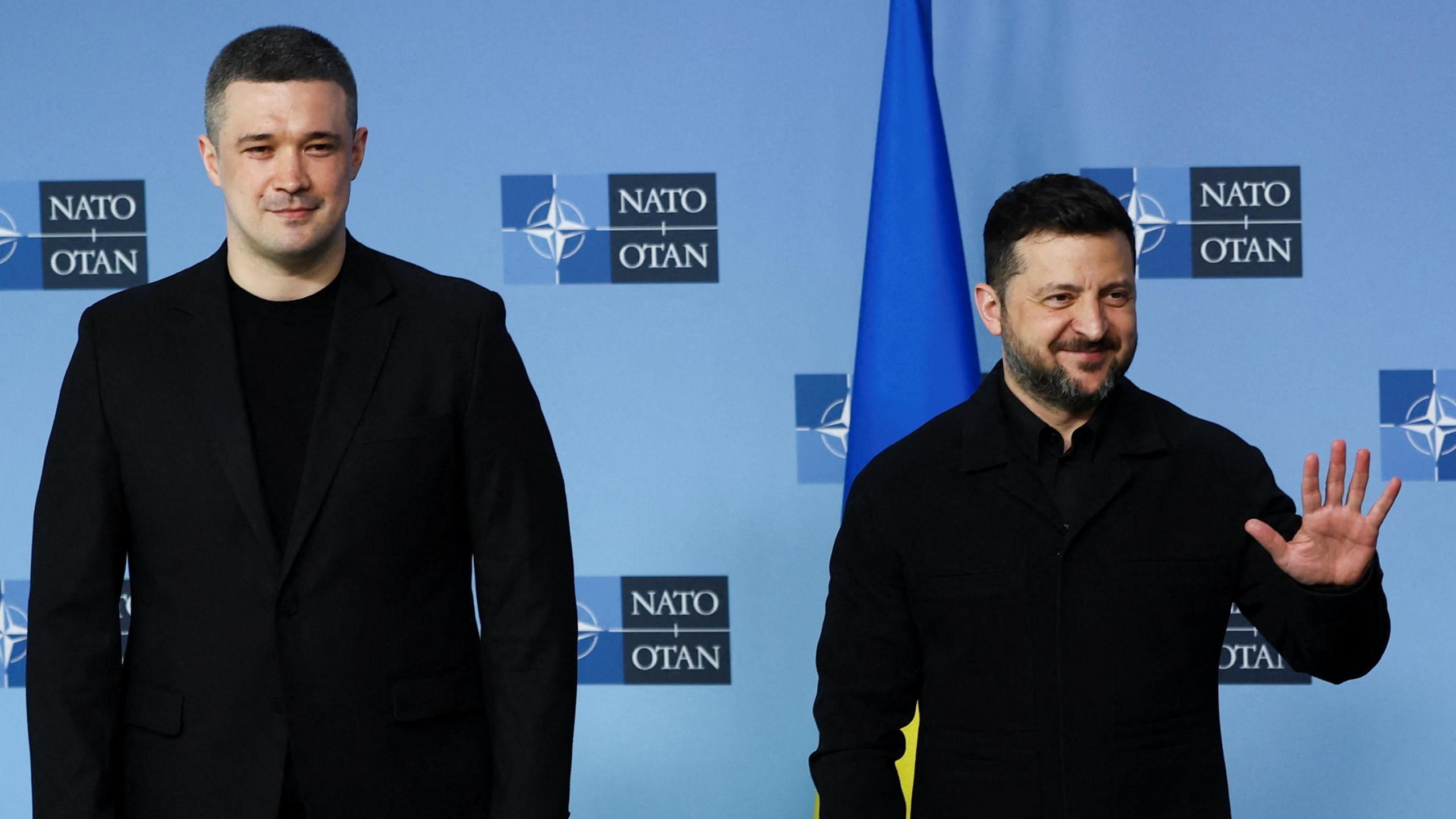 President Volodymyr Zelensky standing smiling with his hand in a hi-five next to another politician also dressed in black in front of a blue background with the Nato symbol and the Ukrainian flag.