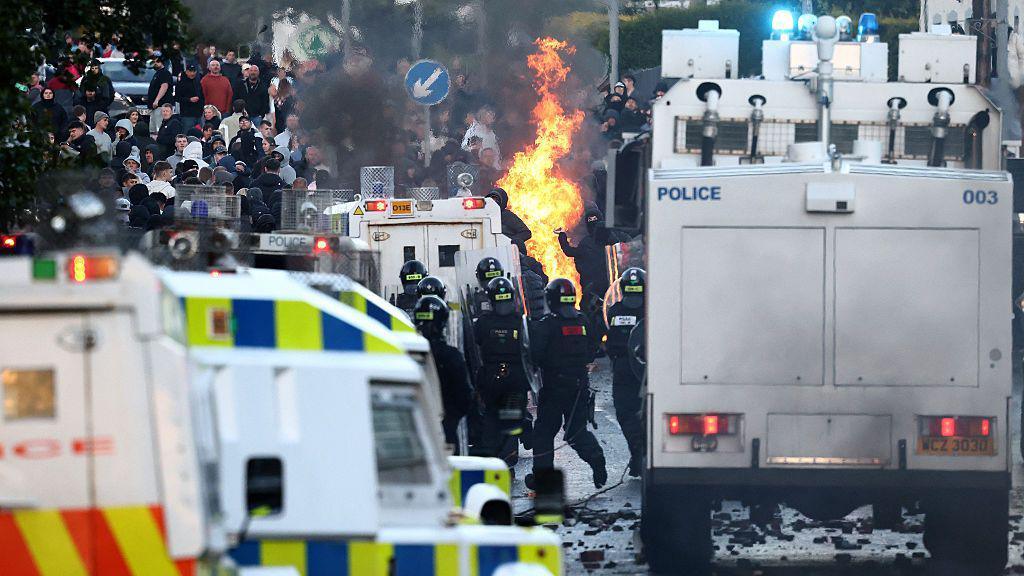 Crowds throw things at Police blocking them from a road. In the foreground are armoured police vehicles and officers in tactical uniforms. In the distance is a large group of people, mostly dressed in dark colours.
