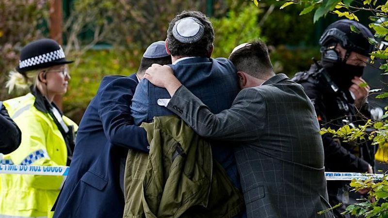 Three men wearing Kippah's hold each other and hug, while on the other side of police tape is a police woman in uniform and a heavily armed police man who is in full black protective gear.