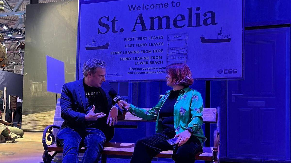 Laura sits with a person wearing a Silent Hill t-shirt and jacket. Behind them, a creepy sign reading Welcome to St. Amelia - a mock-up of something from the game itself.