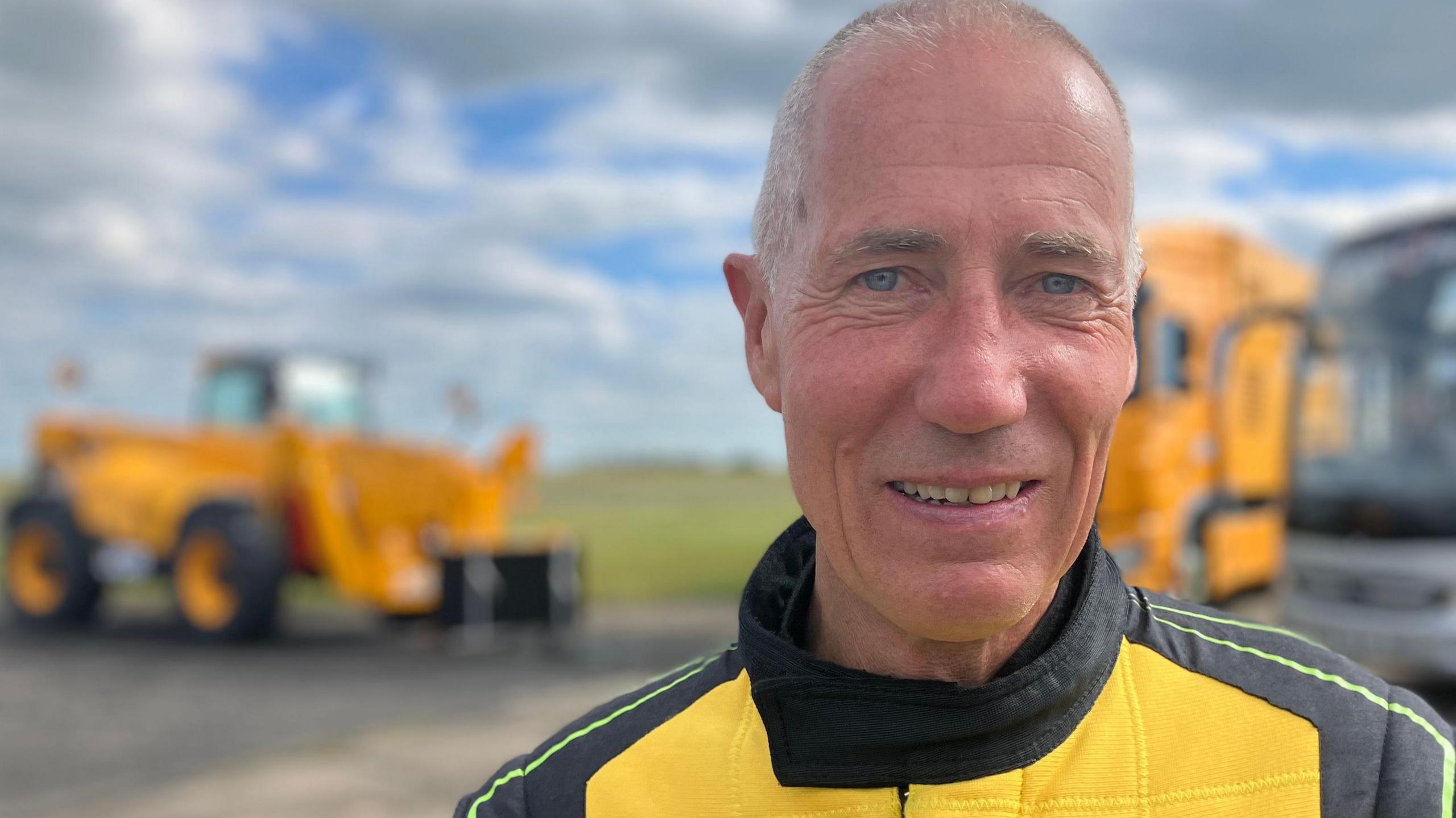 A man looks towards the camera wearing a yellow and black driving suit with the "JCB" logo on the front.