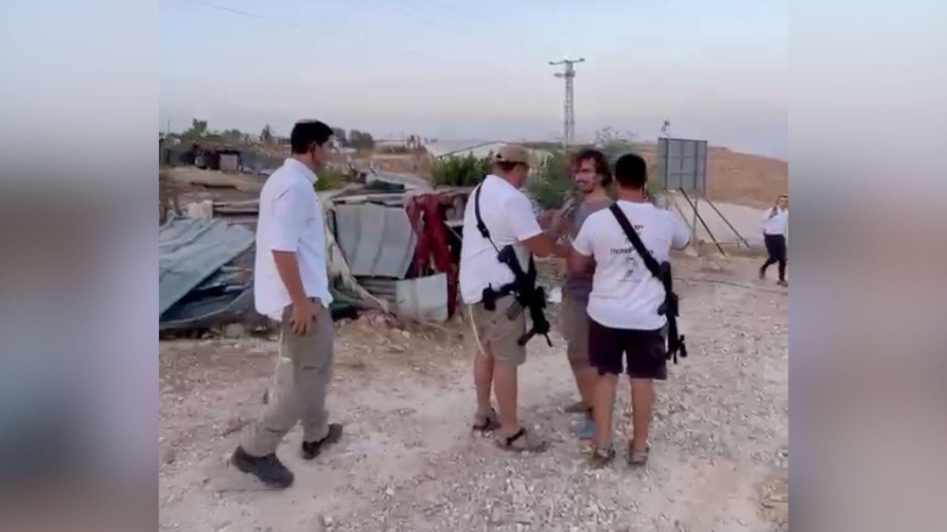 Three Israeli settlers surround a British student in a blurry photo. They wear white T-shirts and carry guns.