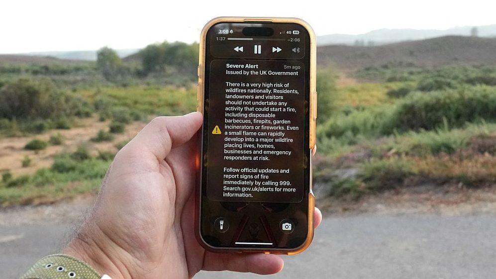 A man holds a phone in front of a green field. The phone is displaying the UK government wildfire alert.