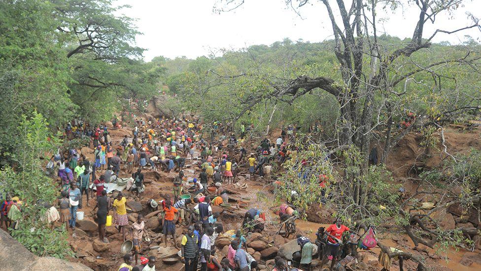 A wide shot showing large numbers of people working on what appears to be a dry river bed. It is surrounded by greenery.