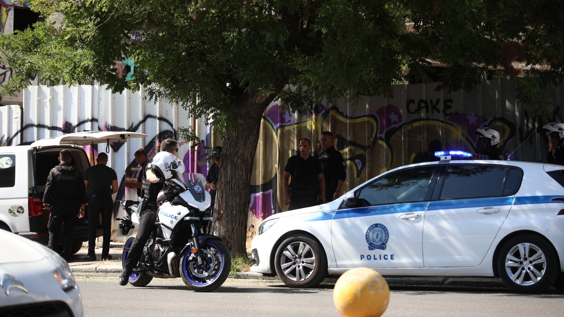 A police care and police officer astride a motorbike in a city street. There is a corrugated fence in the background covered in graffiti.